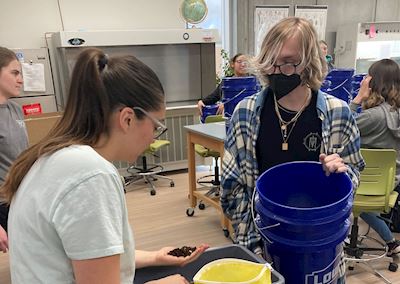 Two students standing over a bucket of dirt on a table. 